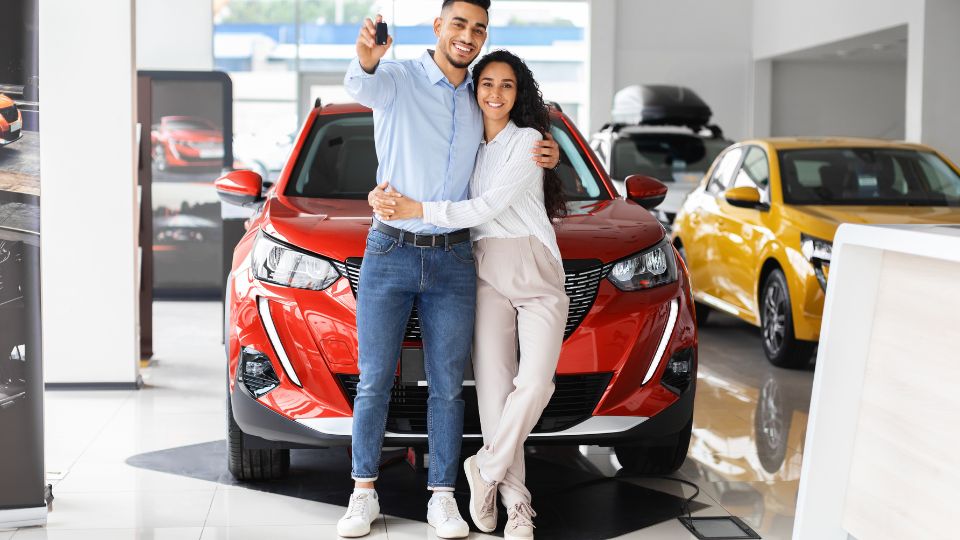 Ghid pentru achiziția unei mașini din străinătate: avantaje, capcane și procesul de înmatriculare Un cuplu fericit stând în fața unei mașini roșii într-un showroom auto.