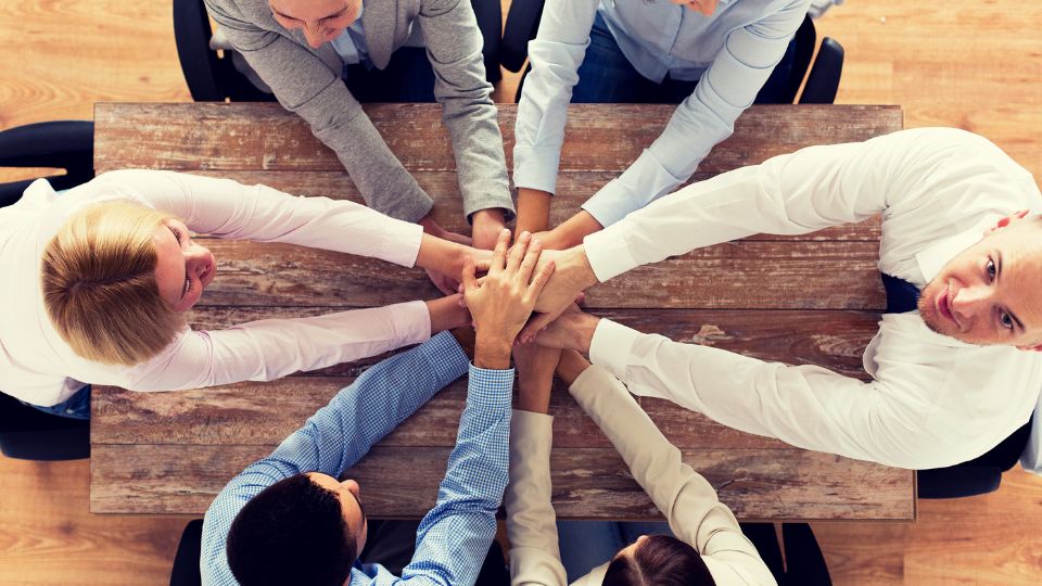 Un grup de oameni își unesc mâinile pe o masă, simbolizând colaborarea.