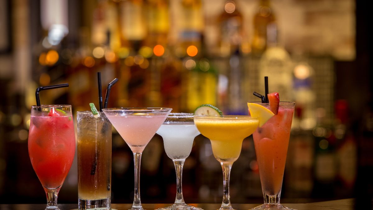 Șase cocktailuri colorate, servite în pahare elegante, pe un fundal de bar.