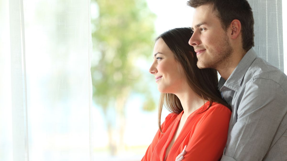Un cuplu zâmbitor stând împreună lângă fereastră, reflectând asupra relației lor.