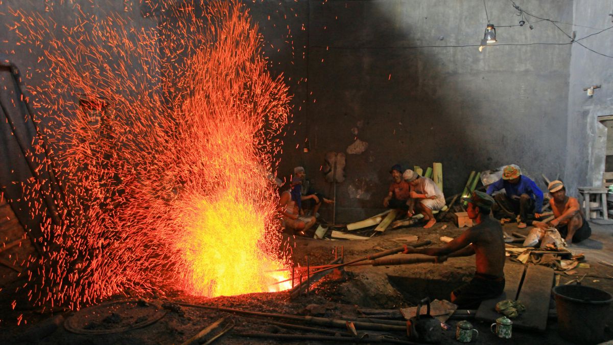 Oameni lucrând într-un atelier de prelucrare a metalelor, cu flăcări și scântei.