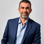 Bărbat cu barbă, îmbrăcat în costum elegant, zâmbind în fața unui fundal deschis.