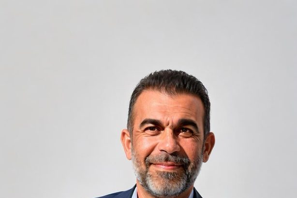 Bărbat cu barbă, îmbrăcat în costum elegant, zâmbind în fața unui fundal deschis.
